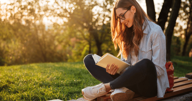 chica sonriente con camisa y anteojos leyendo un libro en una plaza al atardecer