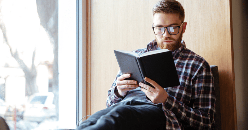 chico con anteojos y camisa leyendo un libro en inglés al lado de una ventana