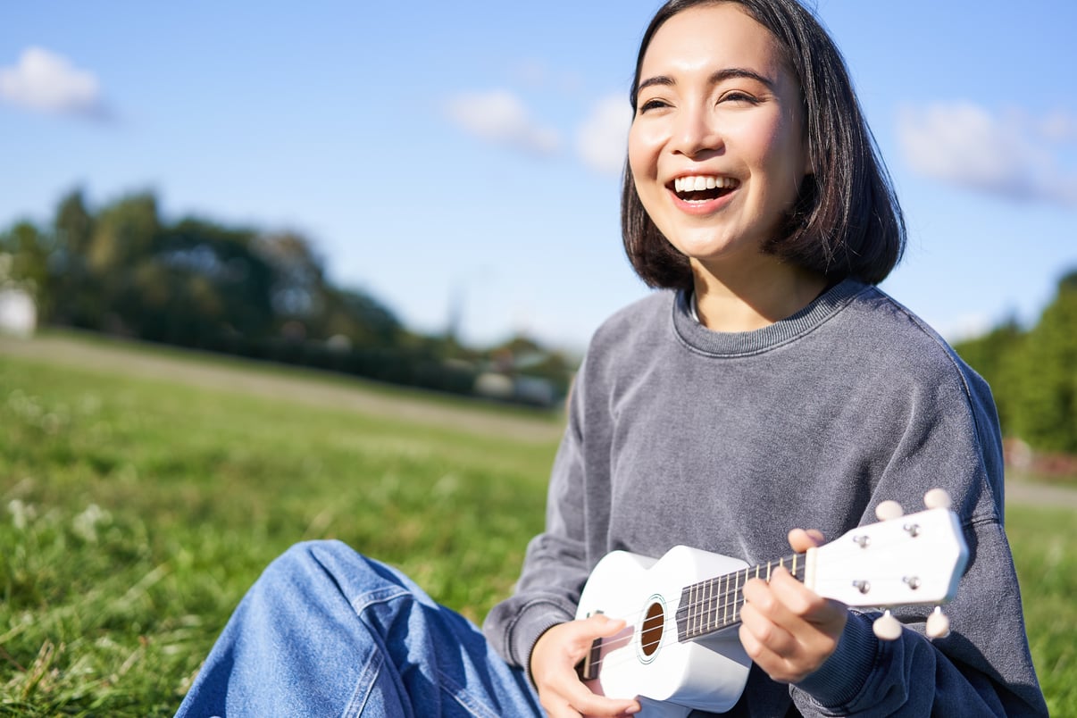 gente-feliz-pasatiempos-nina-asiatica-sonriente-tocando-guitarra-ukelele-cantando-sentado-parque-aventajar