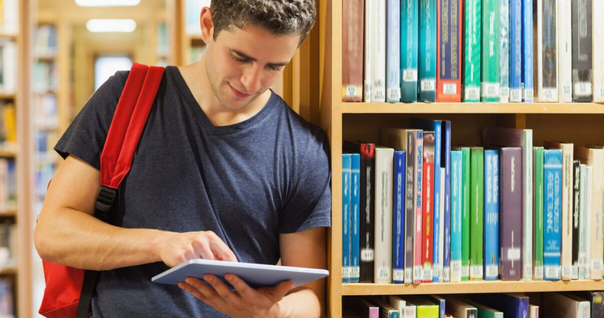 hombre joven en remera y con mochila sosteniendo tablet para aprender inglés en biblioteca de la universidad